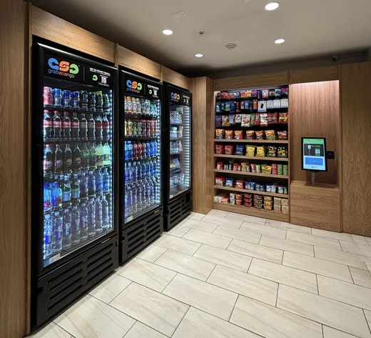 Hotel lobby market with refrigerated drinks, snack shelving and a wall-mounted checkout terminal.