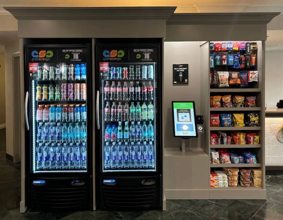 Hotel lobby market with two beverage coolers, snack shelves and a self-checkout terminal.